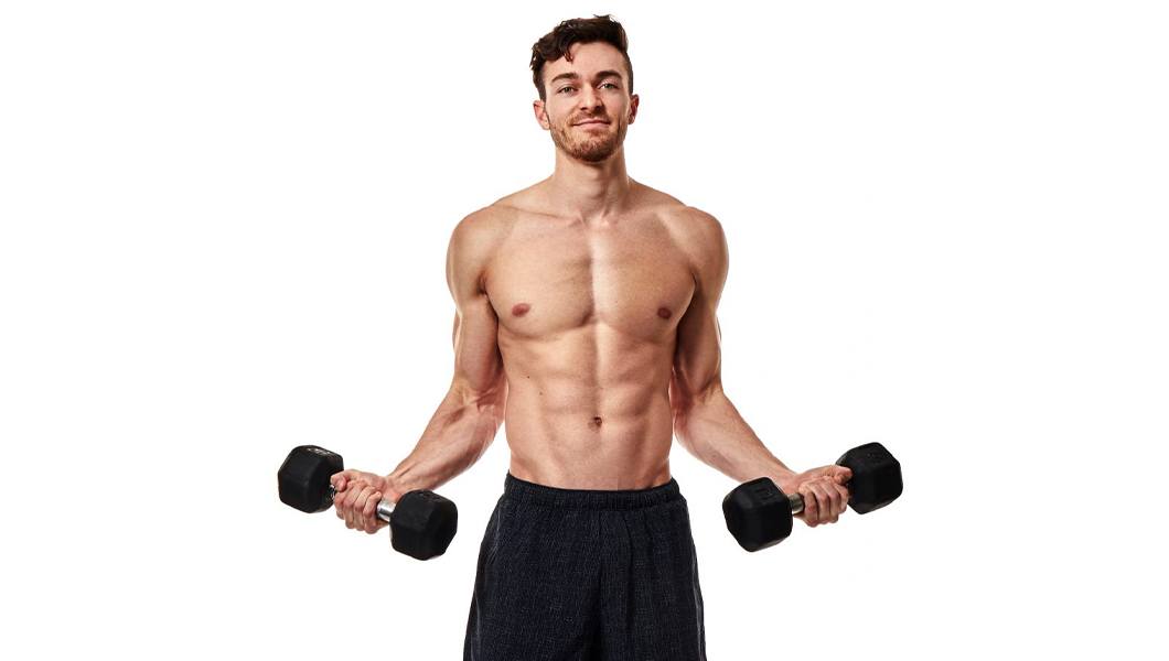 Shirtless Barry's Bootcamp instructor Austin Cagley holding dumbbells by sides on white background