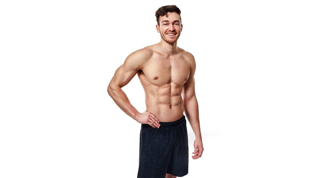 Shirtless Barry's Bootcamp instructor Austin Cagley smiling with hand on hip on white background