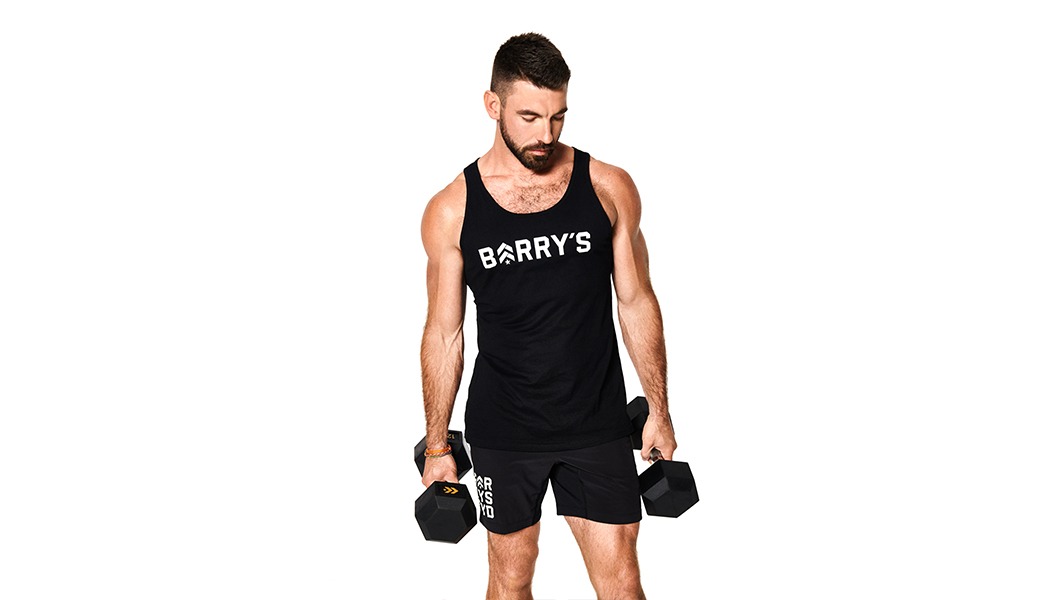 Barry's Bootcamp instructor Cory Gooch wearing a Barry's tank top holding dumbbells and looking down.
