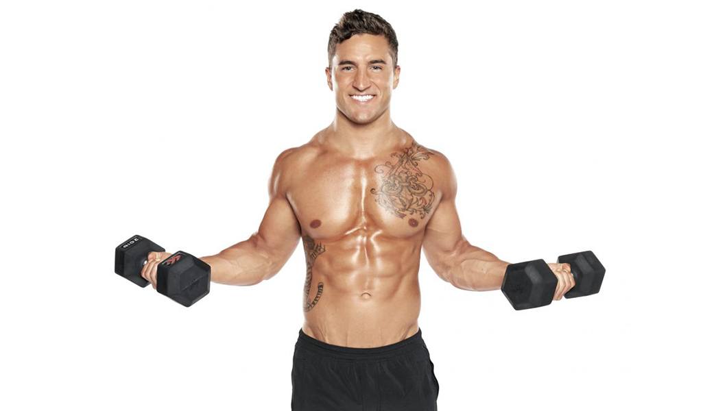 Shirtless Barry's Bootcamp instructor Erick Wilson doing bicep curls with dumbbells and smiling on white background