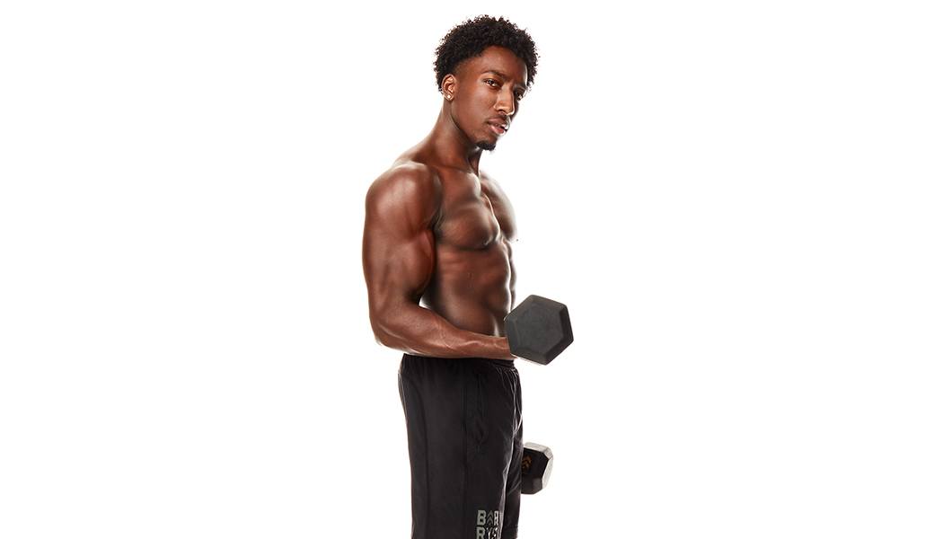 Barry's Bootcamp instructor Joseph Allen shirtless, holding a dumbbell in a bicep curl position.