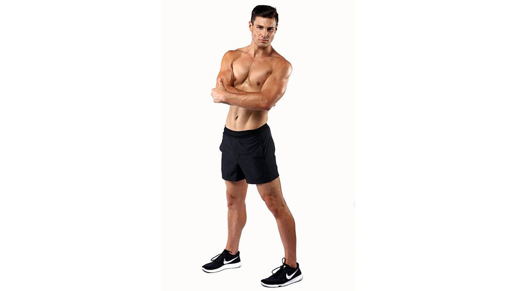 Shirtless Barry's Bootcamp instructor Joe Nicastro smiling with arms folded over chest on white background