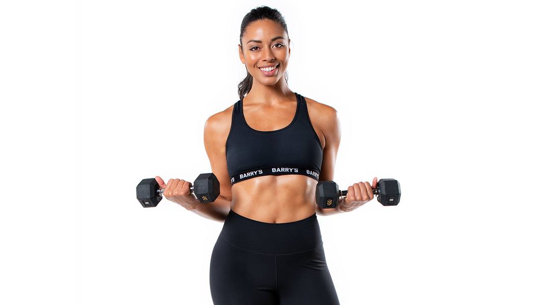 Barry's Bootcamp instructor Mercedes Owens wearing a black sports bra against a white background holding dumbbells out by her side.