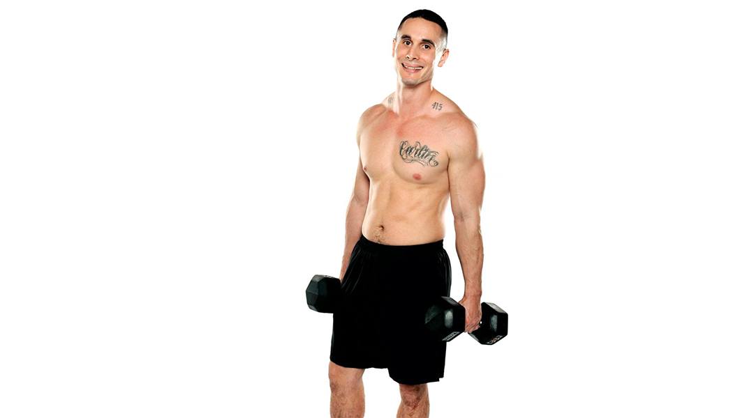 Shirtless Barry's Bootcamp instructor Vidal Carlin smiling and holding dumbbells down by sides on white background