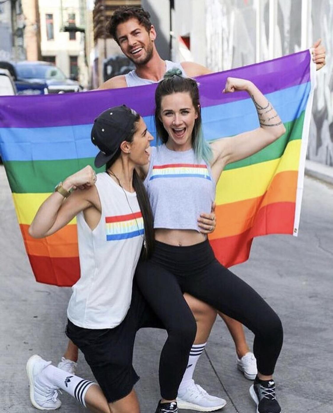 Tres participantes del desfile del orgullo sonriendo y flexionándose frente a la bandera del orgullo del arco iris