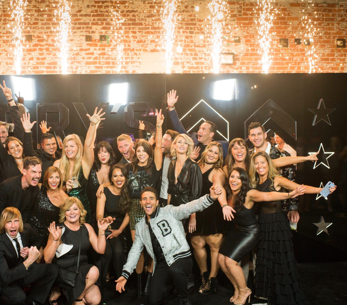 El equipo de Barry's Bootcamp vestido con un atuendo negro semiformal sonriendo con los brazos extendidos frente a la pared con bengalas