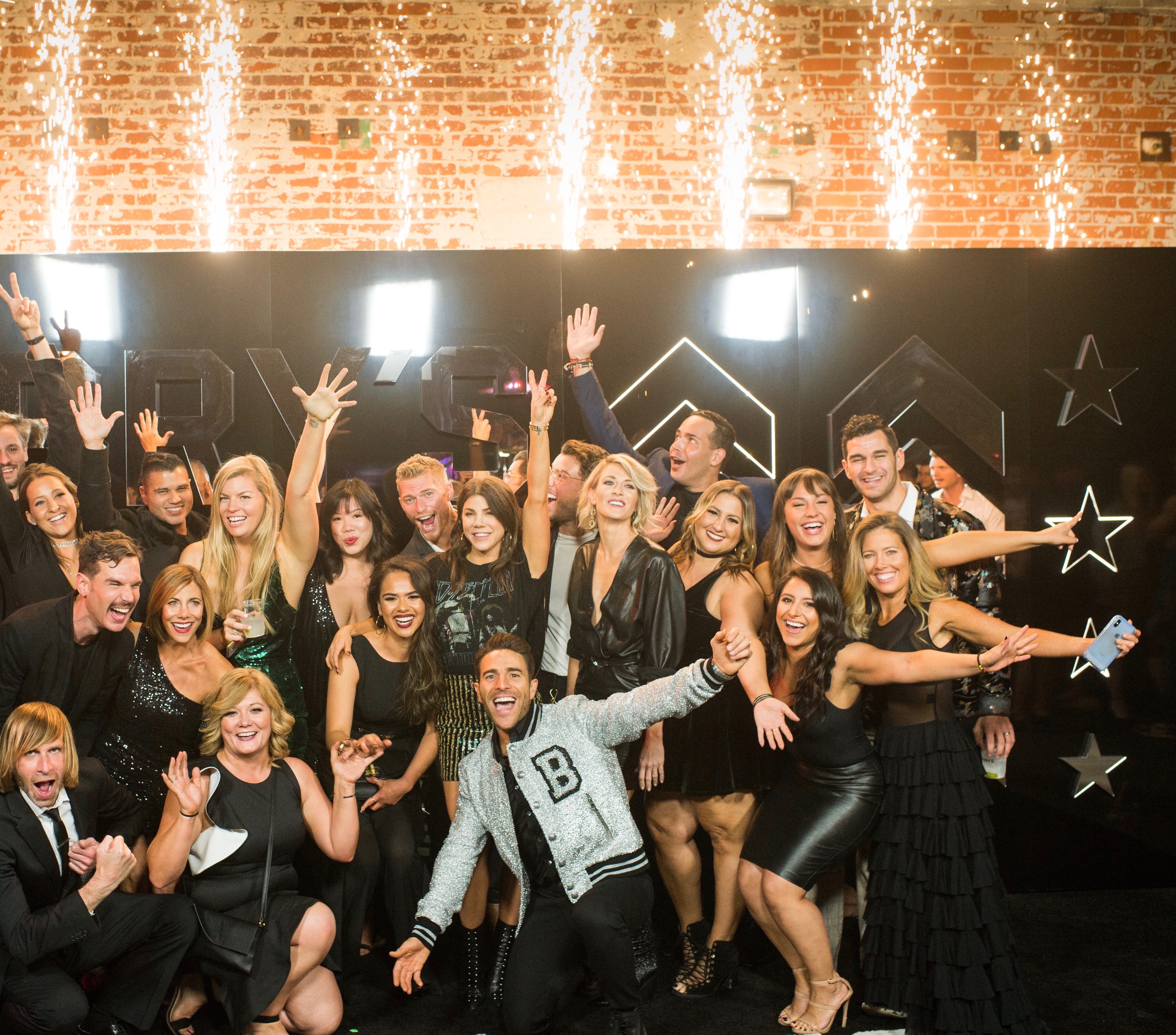 Barry's Bootcamp team dressed in semi-formal black attire smiling with arms outstretched in front of wall with sparklers 
