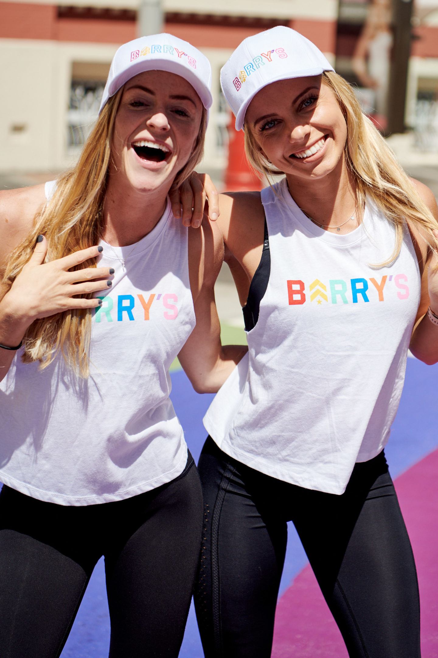 Two young women at Pride parade wearing white tank tops and hats with Barry's printed in rainbow colors