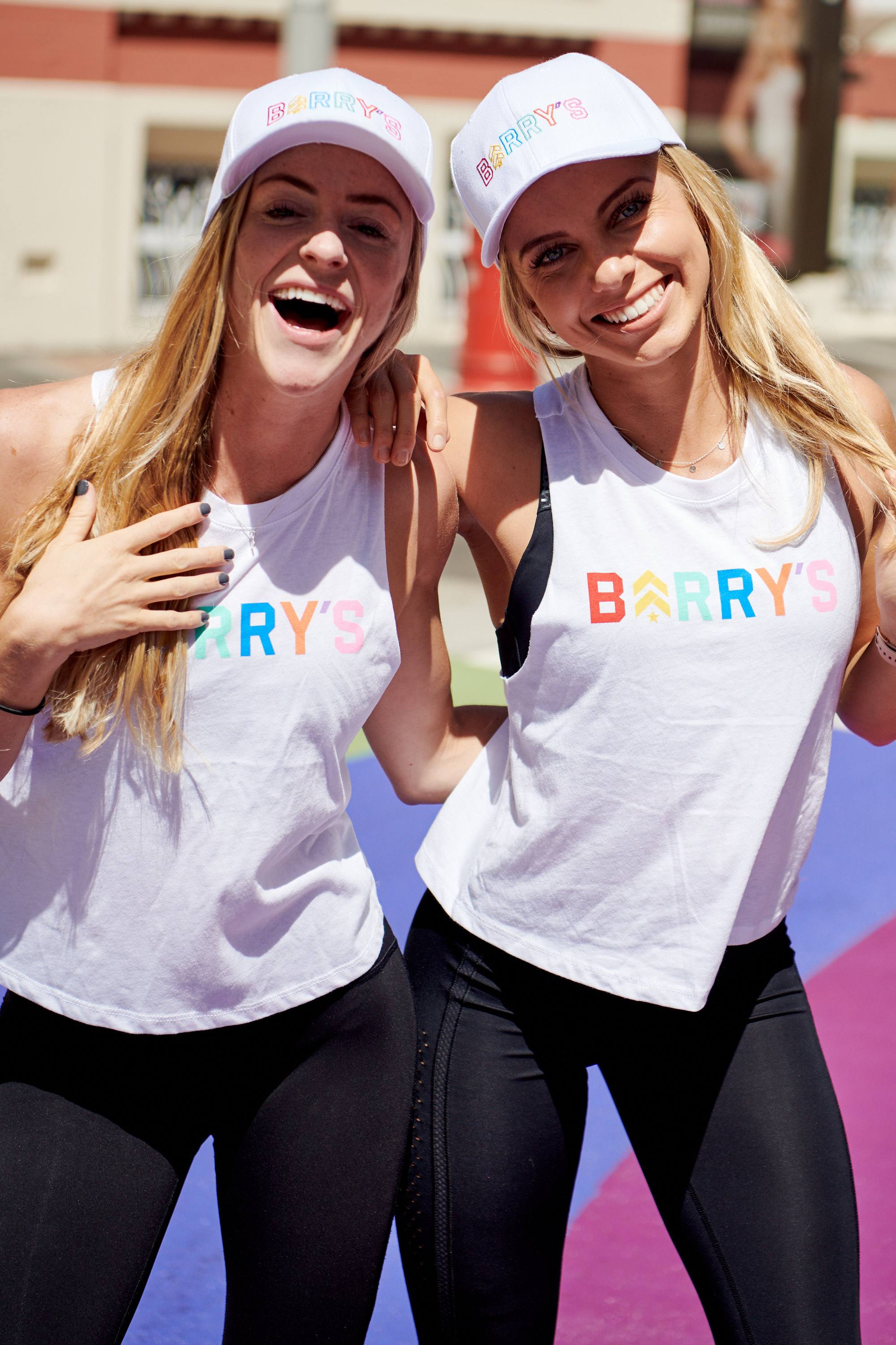 Two young women at Pride parade wearing white tank tops and hats with Barry's printed in rainbow colors 