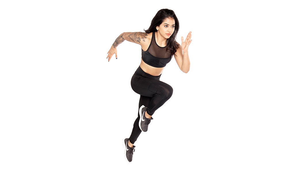 Barry's Bootcamp instructor Trishala Poulose wearing a black sports bra and leggings in a running position against a white background.