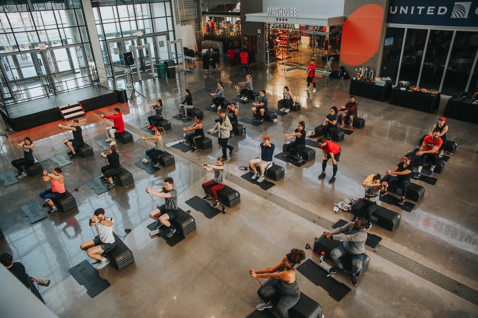 Barry's Bootcamp class in atrium of United Center in Chicago sitting on step up blocks while pulling resistance bands across their chests 