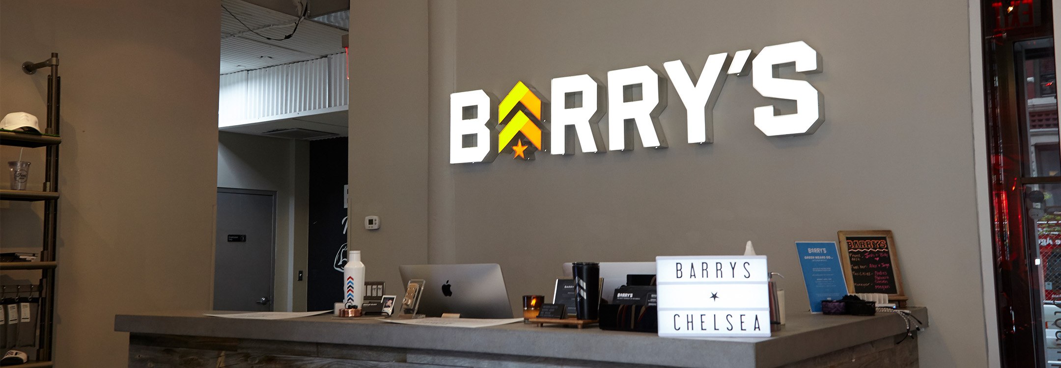 Barry's Bootcamp white sign and yellow and orange logo on wall behind reception desk at Chelsea studio location