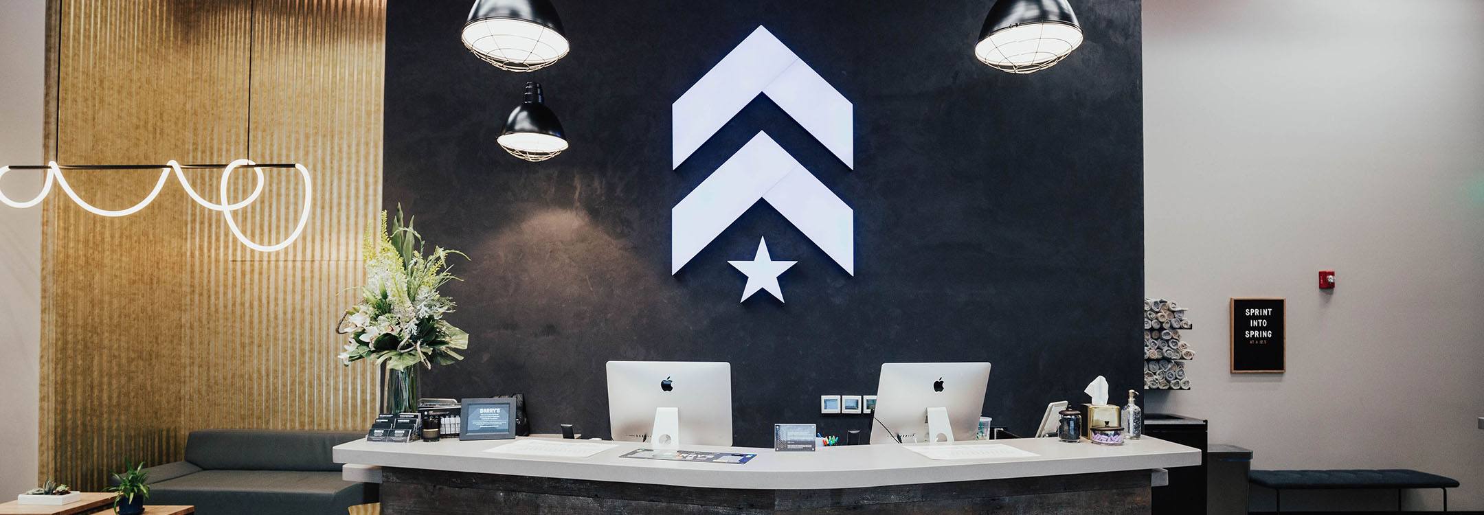 White Barry's Bootcamp upward arrows and star logo on navy wall behind reception desk at FiDi studio location