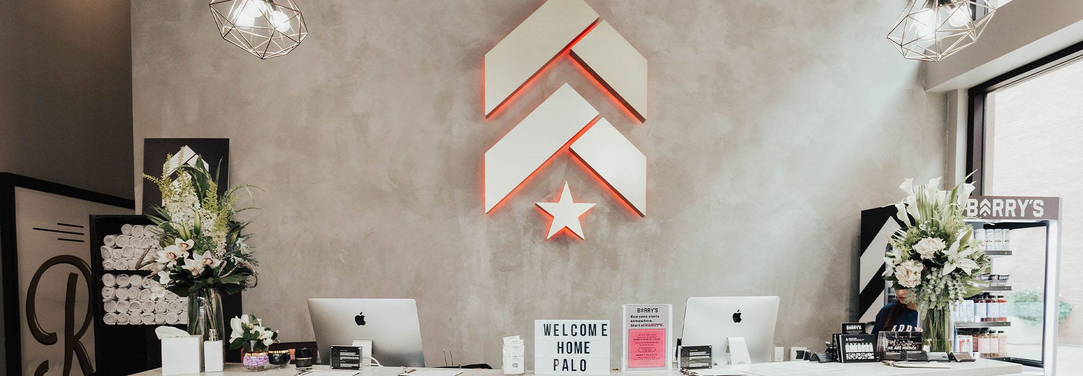 White Barry's Bootcamp upward arrows and star logo with red outline on grey wall in Palo Alto studio reception area