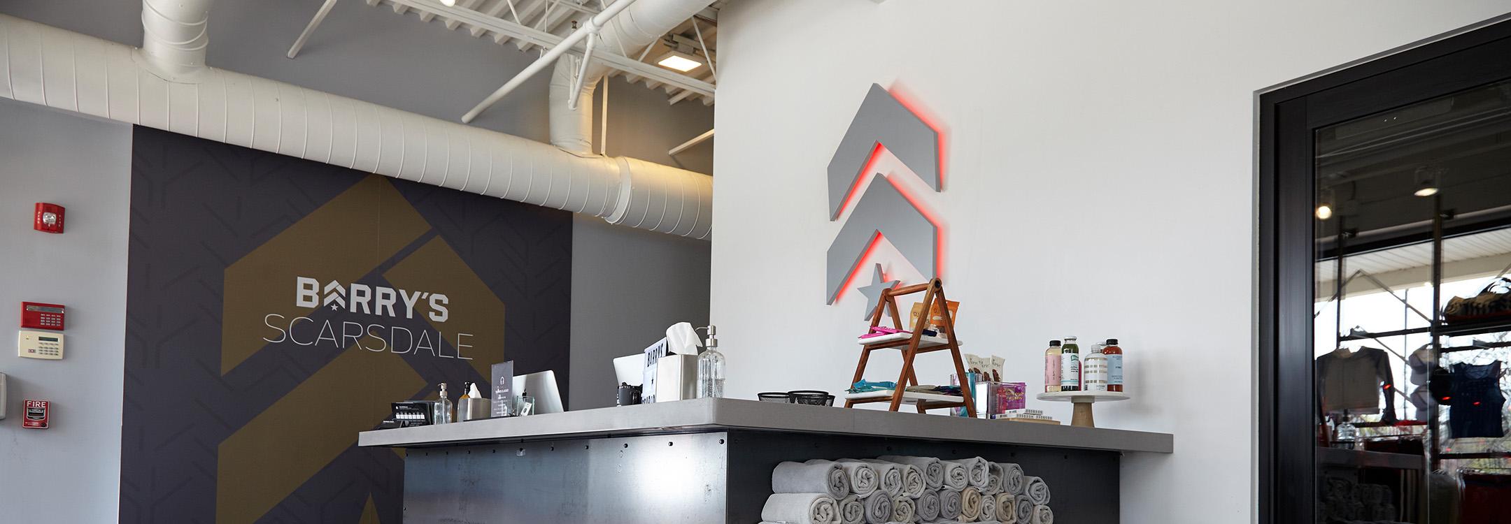 Silver Barry's Bootcamp upward arrows and star logo with red backlighting on wall behind reception desk at Scarsdale studio location