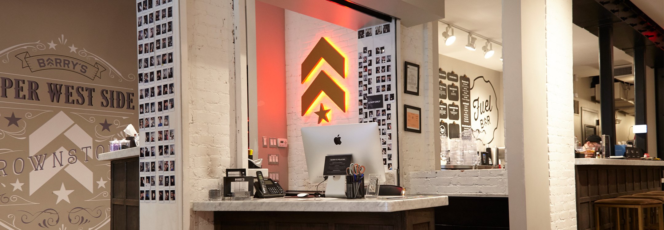 Gold Barry's Bootcamp upward arrows and star logo with red backlighting on white wall behind reception desk at Upper West Side studio location