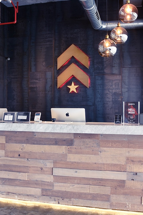 Gold Barry's Bootcamp upward arrows and star logo with red backlighting on grey wall behind reception desk at Dubai Marina studio location