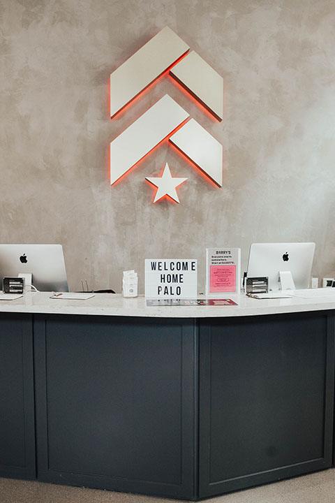 White Barry's Bootcamp upward arrows and star logo with red outline on grey wall in Palo Alto studio reception area