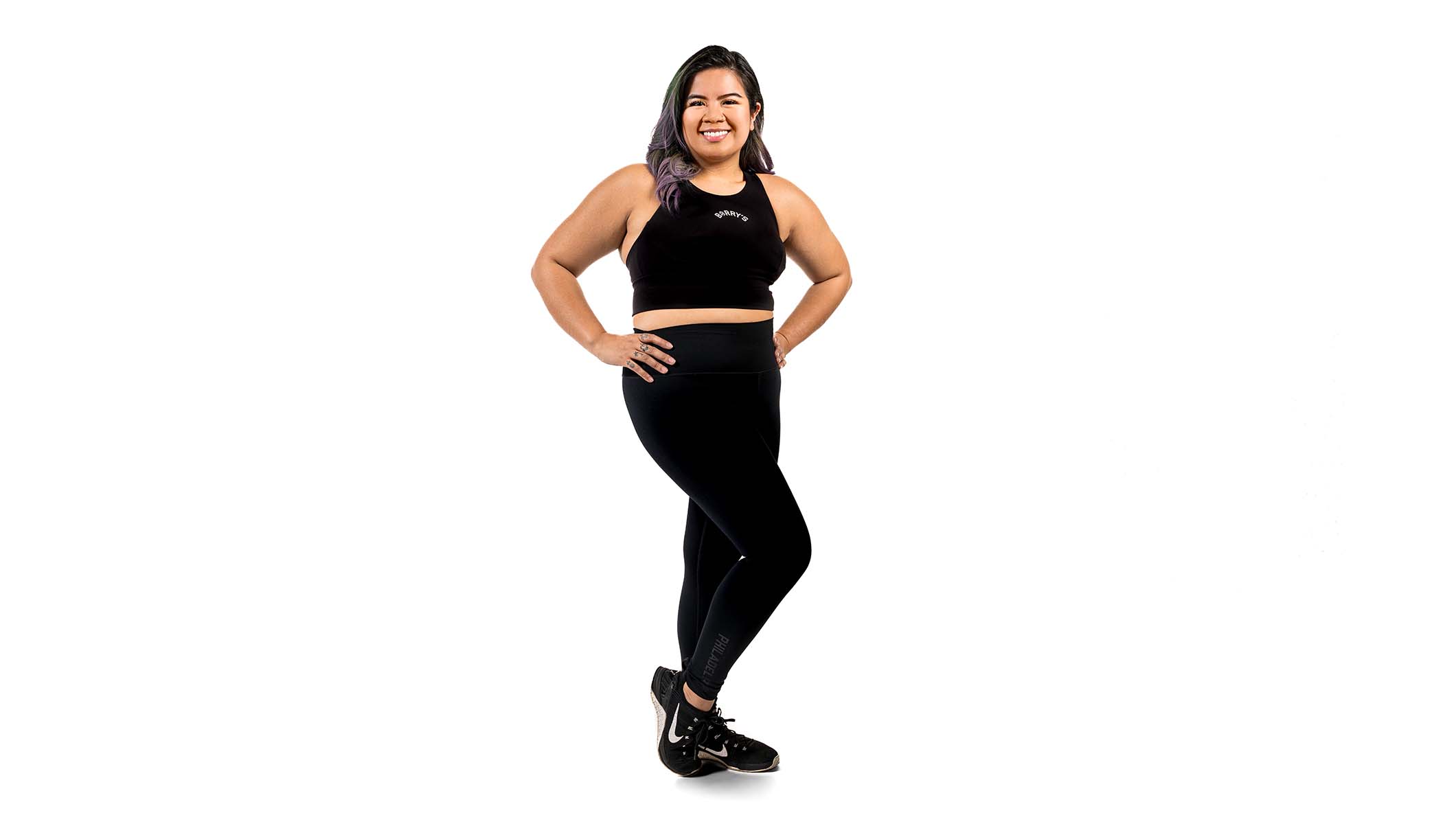 El instructor de Barry, con una camiseta negra, está de pie, sonriendo con confianza sobre un fondo blanco.