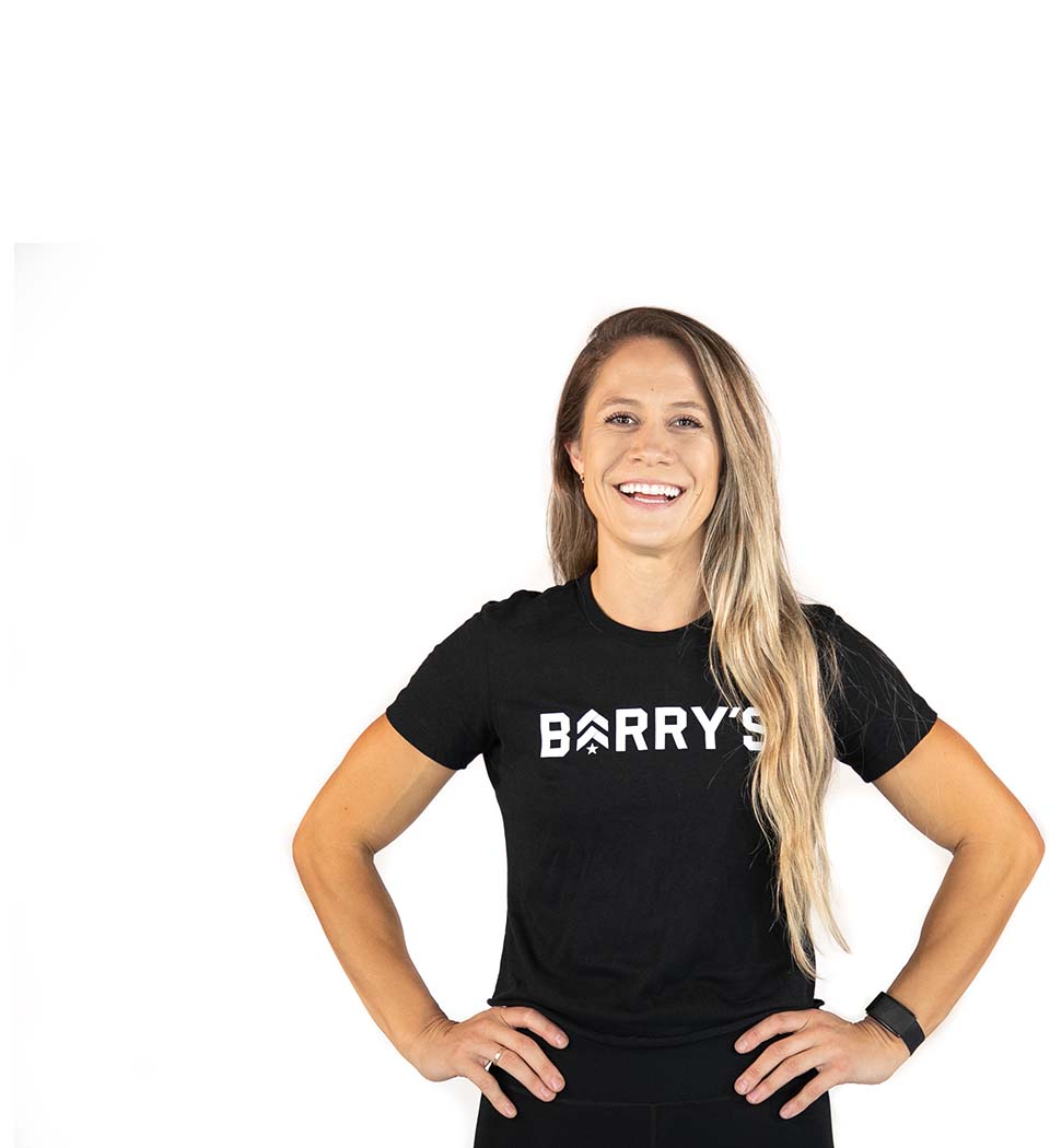 Barry's instructor standing confidently with hands on hips, wearing a black top against a white background, ready for the next workout challenge.