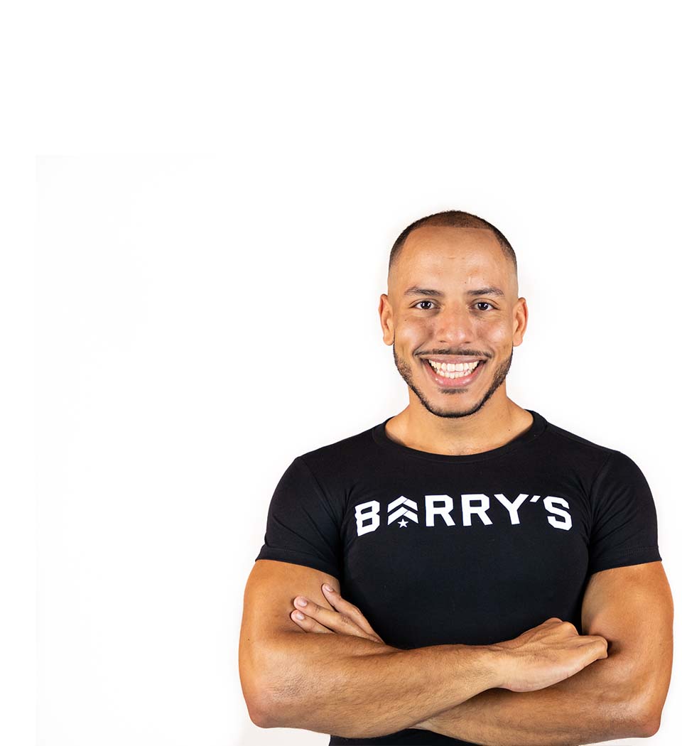 Barry's instructor standing confidently with arms crossed in front, wearing a black tank top against a white background, ready for the next workout challenge.