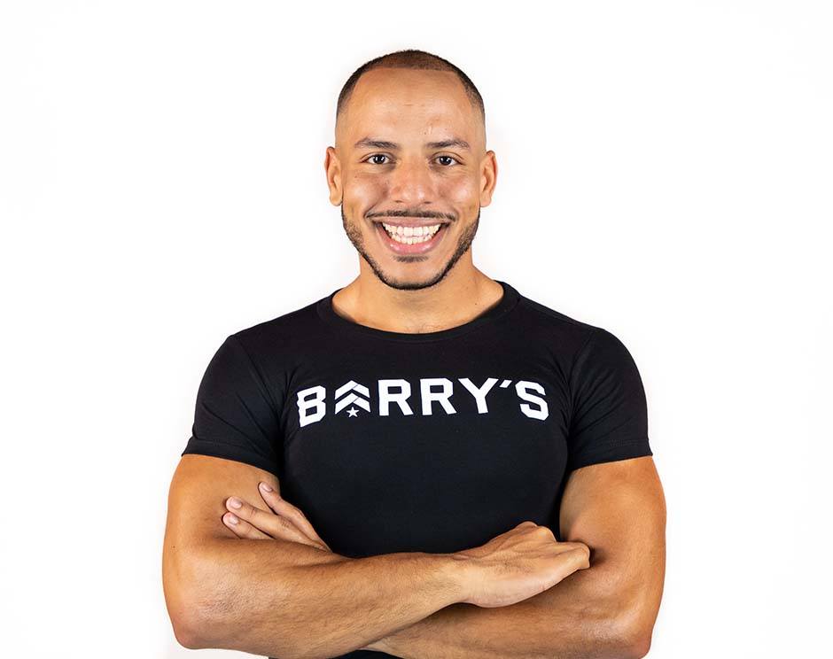 Barry's instructor standing confidently with arms crossed in front, wearing a black tank top against a white background, ready for the next workout challenge.