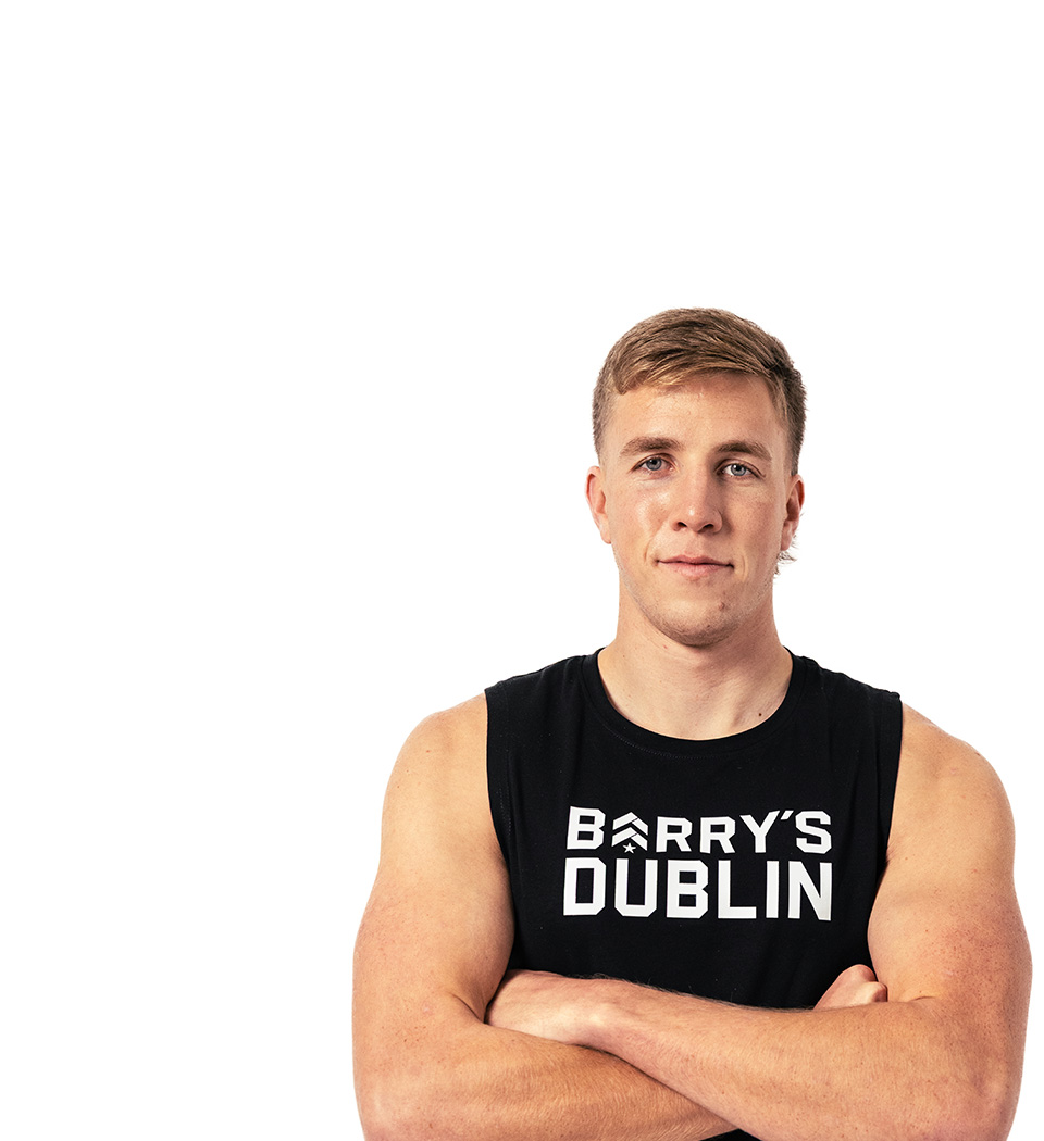 El instructor de Barry, con una camiseta negra, está de pie, sonriendo con confianza sobre un fondo blanco.